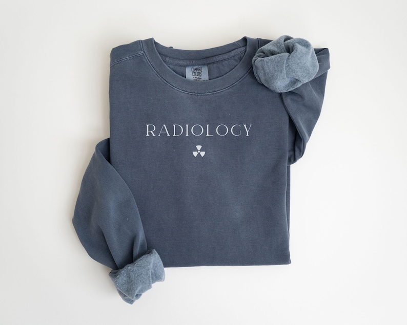 May include: A slate blue sweatshirt with the word "RADIOLOGY" printed in white, along with a radiation symbol. The sleeves are cuffed, and the garment is laid flat on a white surface. The sweatshirt is a Comfort Colors brand.