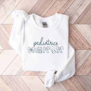 K&ouml;nnte beinhalten: Wei&szlig;er Pullover mit dem Text "P&auml;diatrie" in blauer Schrift. Der Text ist umgeben von kleinen Illustrationen eines Bogens, eines Teddyb&auml;ren, eines Stethoskops, eines Dinosauriers und eines weiteren Bogens.