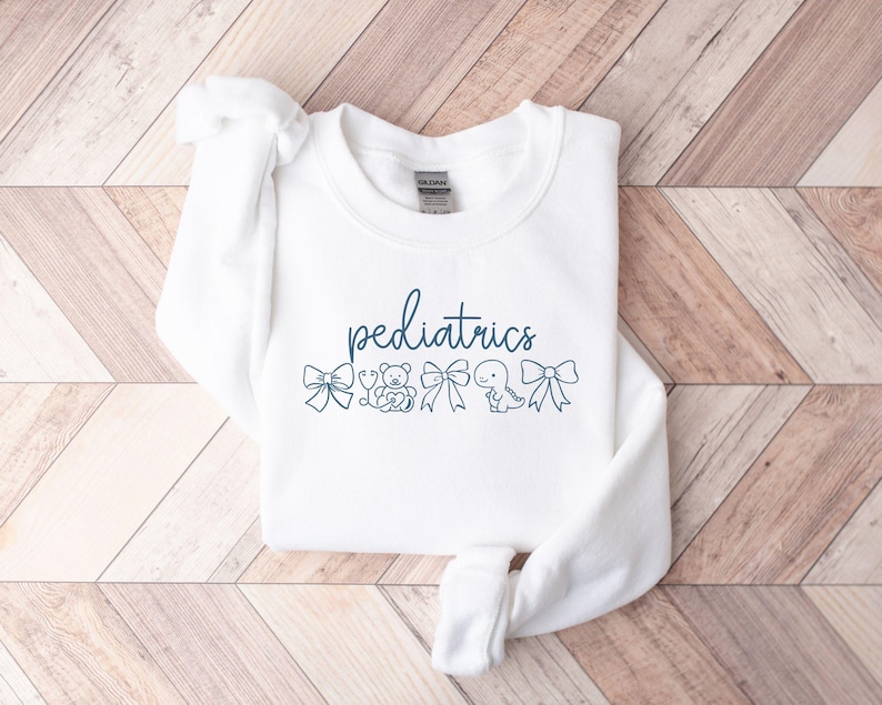K&ouml;nnte beinhalten: Wei&szlig;er Pullover mit dem Text "P&auml;diatrie" in blauer Schrift. Der Text ist umgeben von kleinen Illustrationen eines Bogens, eines Teddyb&auml;ren, eines Stethoskops, eines Dinosauriers und eines weiteren Bogens.