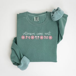 May include: A teal green sweatshirt with the text "intensive care unit" in white script. The text is surrounded by a row of pink flowers with white centers and pink bows.
