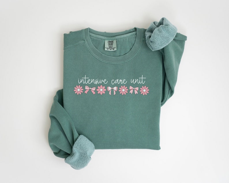 May include: A teal green sweatshirt with the text "intensive care unit" in white script. The text is surrounded by a row of pink flowers with white centers and pink bows.