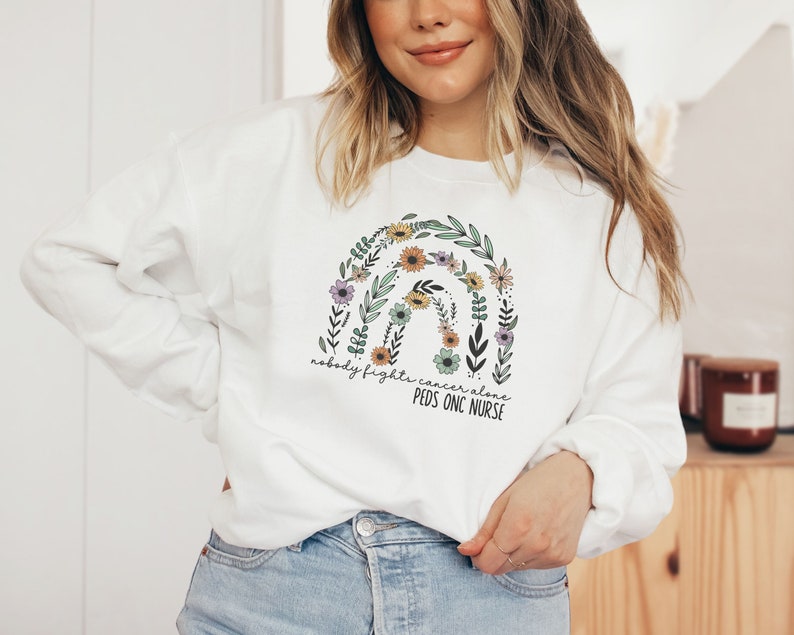 Peut inclure: Sweat-shirt blanc avec un motif arc-en-ciel compos&eacute; de fleurs et de feuilles. Le texte "Nobody fights cancer alone" et "Peds Onc Nurse" est imprim&eacute; sous l'arc-en-ciel.