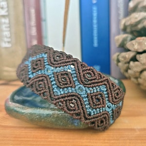 Puede incluir: Un brazalete trenzado marrón y azul con un patrón geométrico. El brazalete está hecho con una técnica de macramé.