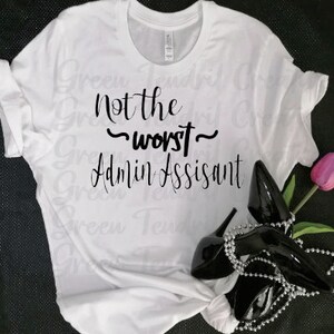 May include: White t-shirt with black text that reads "Not the ~worst~ Admin Assistant". The shirt is lying on a black surface with a pair of black high heels and a pink flower.
