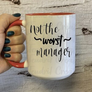 Op de afbeelding: Witte keramische mok met een rode binnenkant en handvat, met de tekst "Not the worst manager" in zwart script. De mok wordt vastgehouden door een hand met donkerblauwe nagellak.