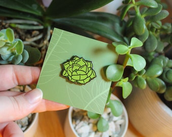 Puede incluir: Un pin esmaltado de suculenta verde lima con un contorno negro, exhibido en una tarjeta verde claro con un patrón de hojas. El pin se sostiene frente a un fondo de plantas verdes y una maceta blanca.