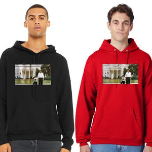 May include: Two black hooded sweatshirts with a graphic print of a man and a child standing in front of a white building with a fence. The sweatshirt on the left is black and the sweatshirt on the right is red.