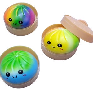 Puede incluir: Tres bollos bao de dibujos animados de colores en cestas de vapor beige. Uno es de color arcoíris, otro amarillo y verde, y el tercero azul y verde. Cada bollo tiene una cara sonriente con ojos negros y una boca curva.