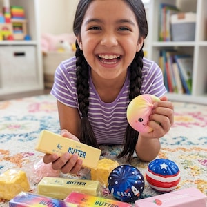 May include: A smiling child holds a rainbow stress ball and toy butter sticks. The toy butter sticks are yellow, rainbow, and pink, with the text "BUTTER" and "4oz" printed on them. Other stress balls include a blue NASA ball and a red, white, and blue ball.