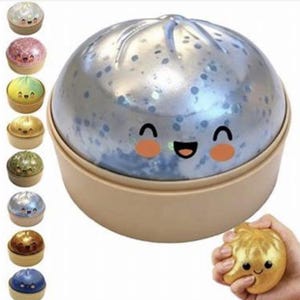 Puede incluir: Una bola antiestrés plateada con forma de dumpling con una cara sonriente, dentro de un recipiente beige. A la izquierda se muestran varias bolas antiestrés más pequeñas y coloridas con forma de dumpling. Una mano aprieta una bola dorada.