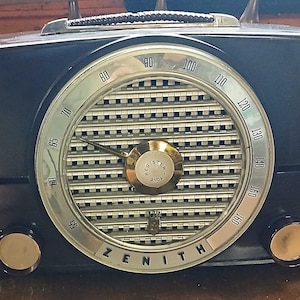 May include: A vintage Zenith tabletop radio with a dark blue rectangular casing, a large circular silver dial featuring frequency numbers from 55 to 160, a central tuning knob labeled Register Tone, and two smaller control knobs on the bottom front.