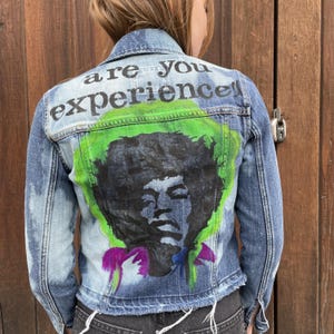 May include: A blue denim jacket with a green, black, and purple painted design on the back. The design features a portrait of Jimi Hendrix and the text "are you experienced?"