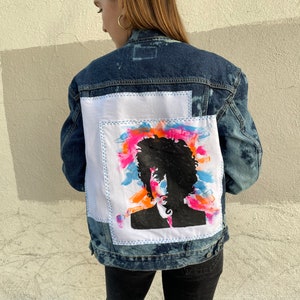 May include: A denim jacket with a white patch sewn on the back. The patch features a colorful abstract design with a black silhouette of a person's head and shoulders.