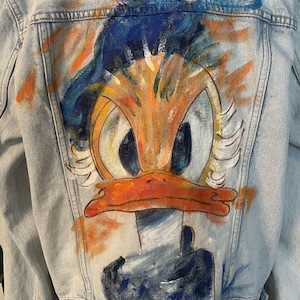 May include: A light blue denim jacket with a hand-painted cartoon image of Donald Duck on the back. The image is in a colorful style with orange, yellow, blue, and white paint.