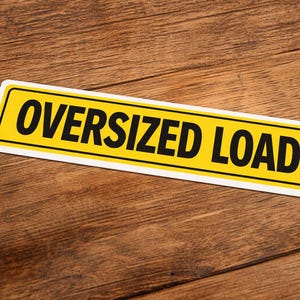 Peut inclure: Panneau rectangulaire jaune avec texte noir « OVERSIZED LOAD ». Le panneau a une bordure noire et un contour blanc. Il est posé sur une surface en bois avec un grain de bois brun.