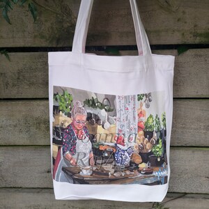 May include: White canvas tote bag with a colorful illustration of a woman in a kitchen. The woman is wearing a red and white patterned shirt and a white apron. She is holding a cup and saucer in her hands. The kitchen is decorated with a white and red floral curtain and a variety of kitchen utensils.