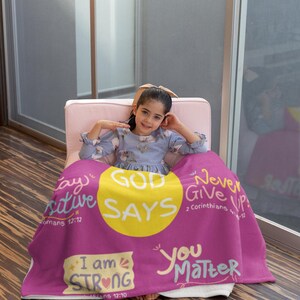 Può includere: Una coperta magenta con testo motivazionale, tra cui "GOD SAYS" in un cerchio giallo, "I am STRONG" e "You Matter". La coperta è drappeggiata su una persona seduta su una sedia rosa. La coperta ha testo bianco ed è realizzata in un materiale morbido.