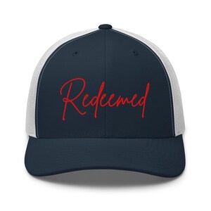 Puede incluir: Gorra de camionero azul marino con parte trasera de malla blanca y visera curva. La palabra "Redeemed" está bordada en rojo en la parte delantera. La gorra tiene un panel frontal estructurado y un cierre a presión ajustable.