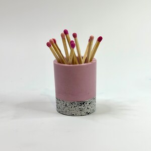 May include: A pink and white concrete matchstick holder with a speckled finish. The holder is filled with wooden matches with pink tips.