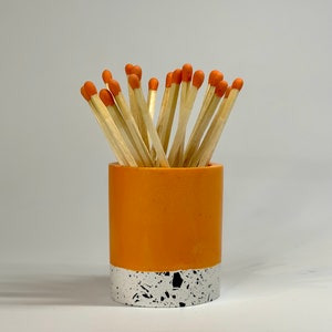 May include: Orange and white ceramic matchstick holder with a speckled pattern. The holder is filled with wooden matches with orange tips.