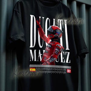 May include: Black t-shirt featuring a graphic of a racer in red racing gear, with the text "DUCATI MARQUEZ" in white. The racer is raising their arm in victory. The number "93" is visible. The shirt has a checkered flag design.