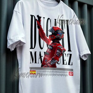May include: White t-shirt featuring a graphic of a racer in a red racing suit and helmet, with the name "DUCATI MARQUEZ" in bold black letters. The shirt includes a checkered flag design and the number 93.