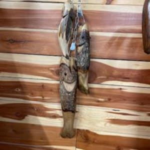 May include: Hand-carved wooden fish hanging from a chain. The fish have a natural wood finish with visible bark and detailed carving. The background is a wood-paneled wall.