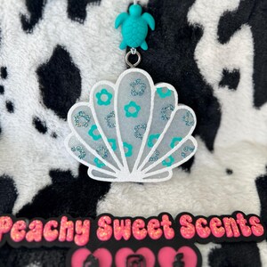 May include: A white seashell-shaped air freshener featuring teal flower accents and silver glitter details, suspended from a teal turtle bead. Below, a black sign displays the text Peachy Sweet Scents with three pink heart icons containing a shop, Instagram, and Facebook logo.