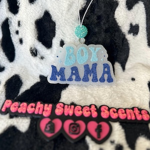May include: A clear, glittery air freshener with the words "BOY MAMA" in blue. The air freshener is hanging from a white string with a turquoise bead. The background is a black and white cow print.