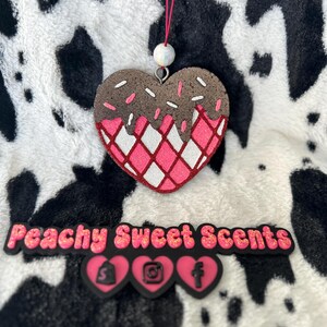 May include: A heart-shaped hanging ornament featuring a brown chocolate-dipped top with white sprinkles and a pink and white waffle-patterned base. Below, a black sign reads Peachy Sweet Scents with three pink heart icons containing black social media logos.