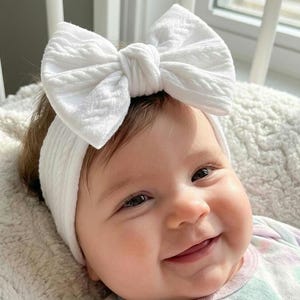 May include: A white cable knit headband featuring a large, matching fabric bow on the front. The accessory is made of a soft, textured material with a classic braided pattern, designed to fit comfortably around the head with a wide, flexible band.
