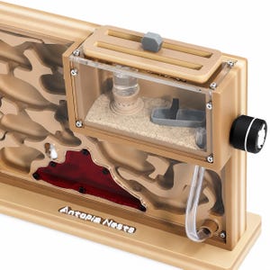 May include: A tan and clear ant farm with the text "Antopia Nests". The ant farm has a clear viewing area, a sand-filled chamber, and a red-tinted area. It features a feeding area and a water reservoir. A black and white knob is on the side.