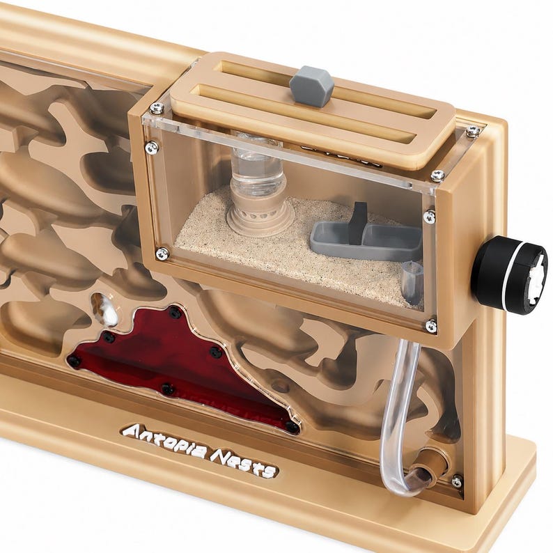 May include: A tan and clear ant farm with the text "Antopia Nests". The ant farm has a clear viewing area, a sand-filled chamber, and a red-tinted area. It features a feeding area and a water reservoir. A black and white knob is on the side.