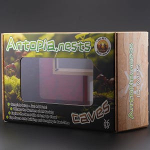 May include: A cardboard box for Antopia nests, featuring a clear window to view the interior. The box has the words "Antopia nests" and "Caves" in a green and white font. The box also has a picture of ants and moss.