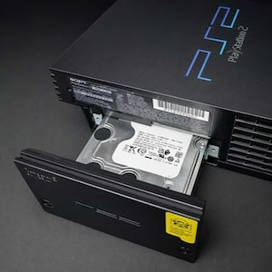 May include: Black PlayStation 2 console with the hard drive bay open, revealing a silver hard drive. The console has the PlayStation 2 logo in blue on the top. The hard drive is partially pulled out.