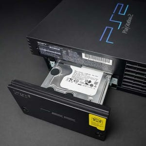 May include: Black PlayStation 2 console with the hard drive bay open, revealing a silver hard drive. The console has the PlayStation 2 logo in blue on the top. The hard drive is partially inserted.