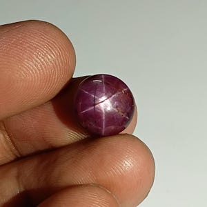 Könnte beinhalten: Ein Rubin-Edelstein im Cabochon-Schliff mit Sterneneffekt. Der Edelstein hat eine tiefe, satte rote Farbe mit einem sechsstrahligen Sternmuster auf seiner Oberfläche. Der Stein ist glatt und poliert, mit einer abgerundeten Form.