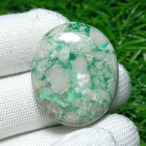 Puede incluir: Gema ovalada con un diseño de mosaico en tonos blancos, verdes y grises. La superficie pulida refleja la luz, mostrando las variaciones naturales de color y textura. Probablemente para joyería o fines decorativos.