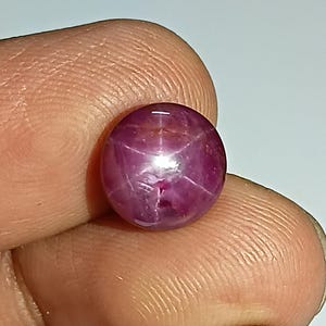 Könnte beinhalten: Ein runder, polierter Rubin-Edelstein mit Sterneneffekt. Der Edelstein hat eine tiefrote bis violette Farbe, mit einem sechsstrahligen Sternmuster, das über seine Oberfläche sichtbar ist. Der Stein wird zwischen Fingern gehalten, um seine Größe und Form zu zeigen.