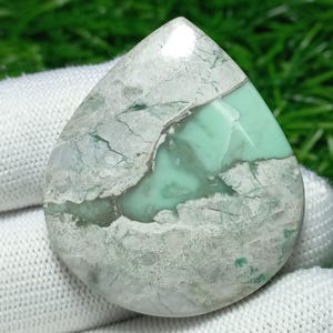 Puede incluir: Una gema en forma de lágrima con una mezcla de tonos verde claro, gris y marrón. La piedra tiene una superficie pulida con vetas y patrones naturales. La gema está sobre un fondo de tela blanca.