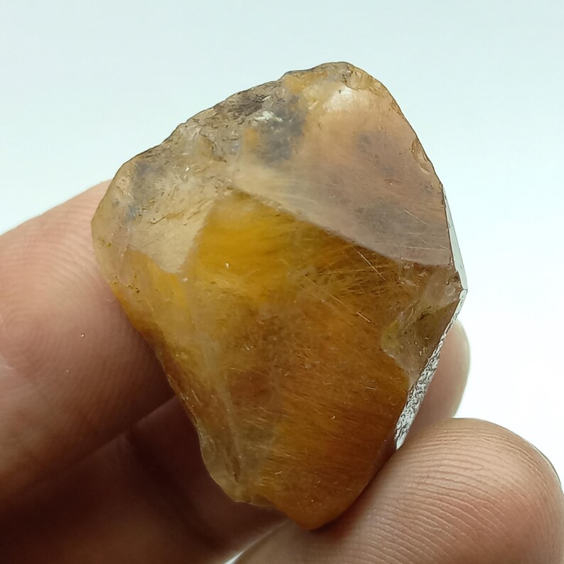 May include: A raw, golden-brown gemstone with a crystalline structure. The stone is translucent, with visible internal inclusions and a faceted edge. The overall shape is irregular, with a natural, unpolished appearance.