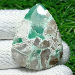 Puede incluir: Una gema en forma de lágrima con una mezcla de colores verde, marrón y blanco. La piedra tiene una superficie pulida con un patrón similar a un mosaico. La gema se utiliza probablemente para joyería o fines decorativos.