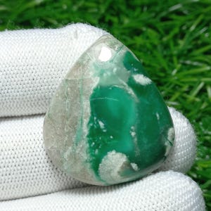 Puede incluir: Una piedra preciosa en forma de lágrima con una superficie pulida. La piedra presenta un tono verde vibrante con vetas blancas y grises. La piedra preciosa se sostiene en un guante blanco, con un fondo de hierba verde.