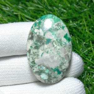 Puede incluir: Una gema ovalada con una mezcla de patrones verdes, blancos y grises. La superficie pulida refleja la luz, resaltando las variaciones naturales de color y textura. La gema se sostiene en una tela blanca.