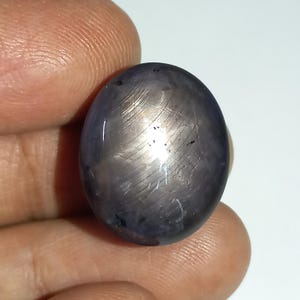 Può includere: Una pietra preziosa a cabochon di forma ovale con una profonda tonalità blu grigiastro e un visibile effetto a stella a sei raggi noto come asterismo. La superficie della pietra presenta una finitura liscia e lucida con inclusioni.