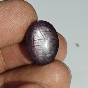 May include: Oval-shaped, purplish-red gemstone with a star-like effect. The polished surface reflects light, showcasing a unique pattern of intersecting lines. The gem is held between fingers, highlighting its size and color.