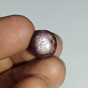 Puede incluir: Una pequeña gema redonda y pulida, de color púrpura intenso. La superficie tiene un brillo metálico reflectante con estrías internas visibles. La gema se sostiene entre dos dedos, mostrando su tamaño y textura.