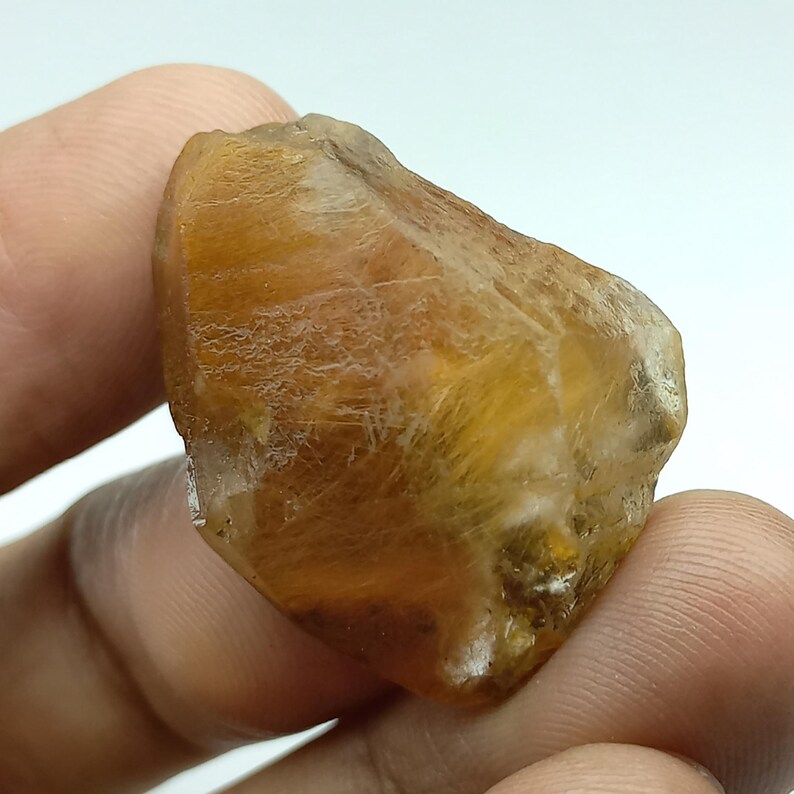 May include: A raw, unpolished gemstone in shades of amber and brown. The surface has a textured appearance with variations in tone. The stone is held in a hand, highlighting its natural, irregular shape and the play of light across its surface.