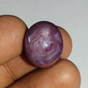 May include: A polished, oval-shaped gemstone with a deep purplish-red hue. The surface displays intricate internal patterns and facets, catching the light. The gem is held between fingers, showcasing its size and shape.