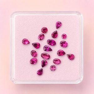 May include: A collection of loose, faceted magenta gemstones in various shapes, including pear, oval, and round cuts, arranged on a light pink velvet insert inside a clear, square plastic display case with a transparent lid.